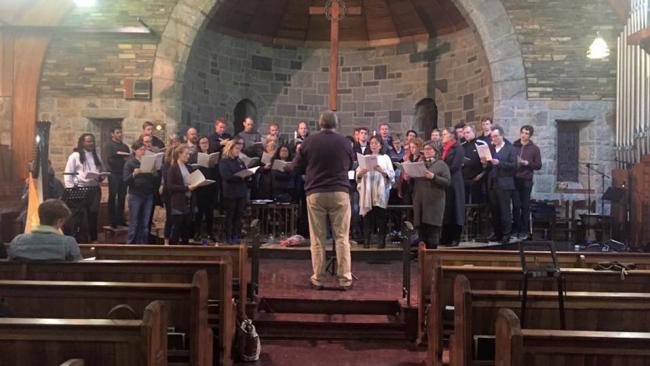Photo of Approx 30 choir members in causal clothes standing in front of a church altar, holding music and being led by a conductor