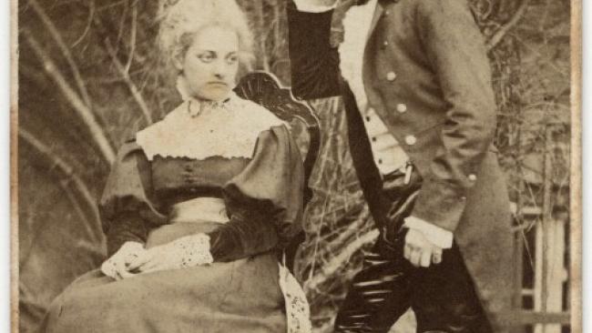 Image of Black and white photo. A sitting woman wearing Victorian dress, a man standing with his elbow leaning on the back of the chair.
