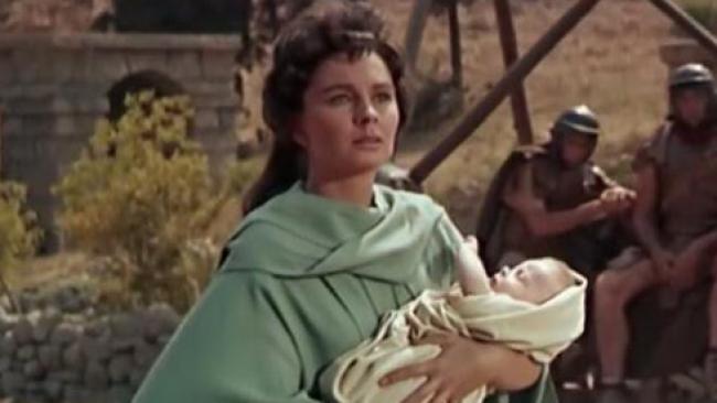 A screenshot of a film. A woman wearing a robe and carrying a baby stands in front of men dressed as Roman soldiers.