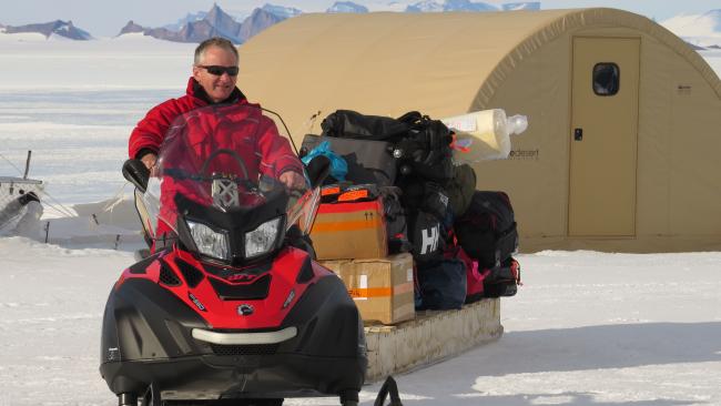 Professor Julian Dowdeswell riding a skidoo and pulling a trailer of luggage across a snowy landscape