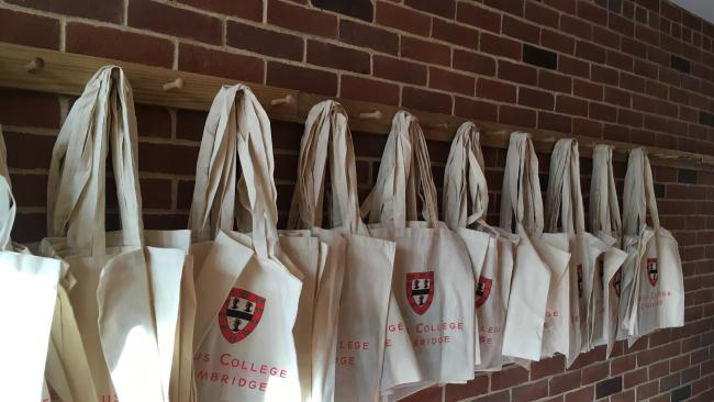 Jesus College tote bags hung on a row of wooden pegs set into a brick wall.